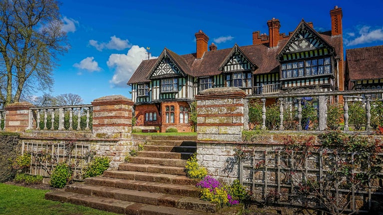 Sanding to the right at the bottom of the steps up to the South Terrace at Wightwick Manor. The half timbered house is in the background behind wooden balustrades where the roses grow in the beds.